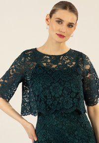 Türkiser Spitzenoberteil mit kurzen Ärmeln, das mit floralen Mustern und einem geschichteten Design versehen ist. Glatte Stoffstruktur mit rundem Ausschnitt.