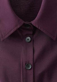 Colletto di camicia viola scuro con una texture liscia, dotato di quattro bottoni neri tondi e di una cucitura dettagliata lungo i bordi.
