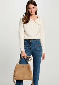 Off-white gerippter Pullover mit weiter Kragen und einem Reißverschluss, kombiniert mit blauen Jeans, die goldene Knöpfe haben, und einer hellbraunen Wildlederhandtasche mit Fransen.