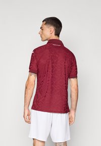 Burgundska polo majica s teksturiranim uzorkom, ovratnik s gumbima i logotipom na leđima. Kratke rukave s kontrastnim obrubom, u kombinaciji s bijelim kratkim hlačama.