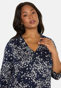 Kukkakuosiset V-pääntiehupparit, joissa on tummansininen tausta ja valkoisia kukkakuoseja, pitkät hihat ja teksturoitu kangas. Koristeltu kultaketjulla.