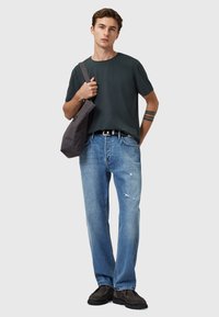 Dunkelgrünes T-Shirt mit kurzen Ärmeln, hellblaue, verwaschene Jeans, graue Schultertasche und schwarze Schuhe. Einfaches, lässiges Outfit mit subtilen Designdetails.