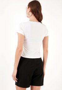 Top bianco a maniche corte con design aderente, abbinato a pantaloni corti neri, che mostra una texture liscia e un taglio leggermente crop.