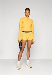 Casaco e calções desportivos amarelos feitos de tecido leve. Modelo segura uma garrafa de água preta, usa meias brancas e ténis claros.