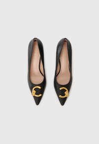 Chaussures à talons hauts en cuir noir avec un bout pointu et un accent en métal doré en forme de cercle sur le dessus. Texture lisse avec un design minimal.