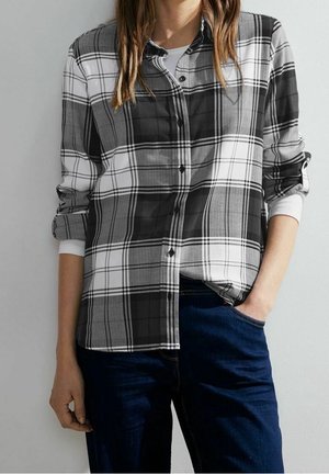 Kvinde iført en sort, hvid og grå ternet skjorte med knapper over en hvid langærmet top, kombineret med mørkeblå jeans, med den ene hånd i lommen.