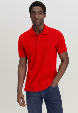 Polo shirt en coton rouge avec un col classique, patte de boutonnage à trois boutons et manches courtes, associé à un jean en denim bleu foncé.