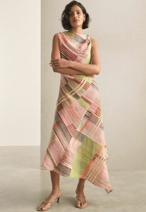 Frau mit kurzen Haaren, die ein ärmelloses Midi-Kleid mit mehrfarbigem geometrischem Muster und beige Sandaletten mit Absatz trägt, steht mit verschränkten Armen.