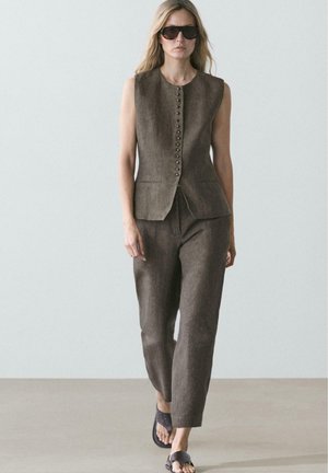 Femme marchant avec un gilet sans manches marron boutonné et un pantalon assorti, portant des lunettes de soleil foncées et des sandales noires à bout ouvert.