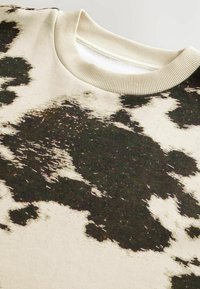 Pull en coton à col ras du cou avec une base beige, présentant un imprimé abstrait de vache en noir et marron. Encolure côtelée et texture douce.