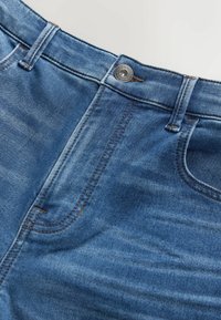 Detailaufnahme von blauen Jeans aus Denim mit Knopf, Gürtelschlaufen, Vordertasche und Naht auf glatter Stoffoberfläche.