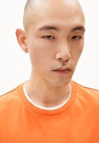 T-shirt en coton orange vif avec un col rond et des manches courtes. Col intérieur blanc visible. Le modèle a la tête rasée et porte une boucle d'oreille.