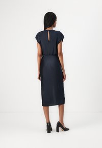 Abito midi plissettato blu navy con maniche a cappuccio e dettaglio a chiave sulla schiena, abbinato a scarpe nere con tacco alto. Tessuto dalla texture liscia.