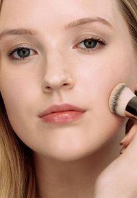 Sminkebørste som påfører foundation på en lys hudfarge; synlig glatt hud, naturlig leppefarge og nøytral øyemakeup; lysebrunt hår.