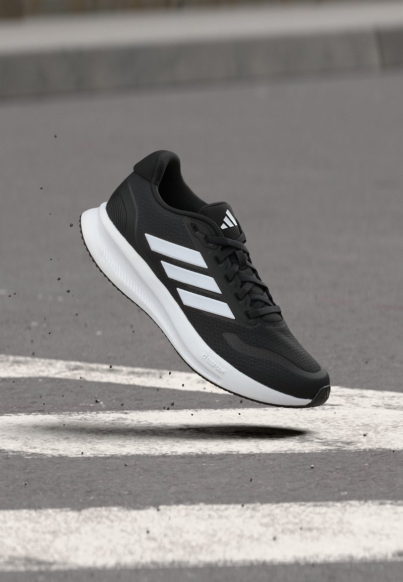 Chaussure de course noire avec semelle blanche et trois bandes blanches, suspendue au-dessus d'une route en asphalte gris avec des lignes blanches de passage piéton.
