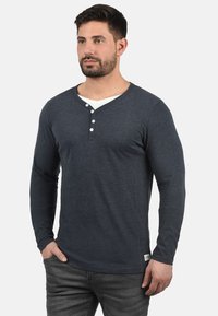 Mørkeblå langærmet henley-skjorte lavet af blødt stof, med V-hals, tre hvide knapper og et lille mærke ved kanten.