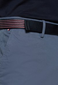 Gros plan sur une personne portant un pantalon bleu et une ceinture en tissu rayée noire, rouge et blanche avec une chemise sombre rentrée.