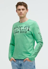 Grünes Langarm-T-Shirt mit Rundhalsausschnitt. Mit schwarzem Drucktext und Designs auf der Vorderseite und dem Ärmel. Getragen mit hellblauen Jeans.