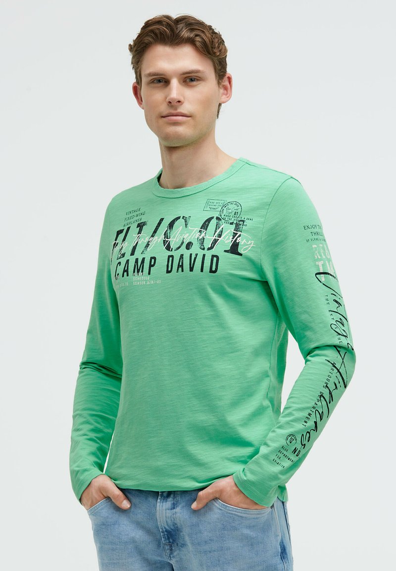 Grünes Langarm-T-Shirt mit Rundhalsausschnitt. Mit schwarzem Drucktext und Designs auf der Vorderseite und dem Ärmel. Getragen mit hellblauen Jeans.
