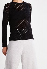 Svart stickad långärmad topp med ett nätmönster och stora hål. Rund halsringning och figurnära midja. Styklad med ljusgrå vida byxor.