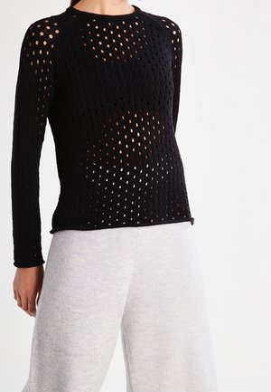Czarny dzianinowy sweter z długim rękawem, charakteryzujący się siateczkowym wzorem z dużymi otworami. Okrągły dekolt i dopasowana talia. W połączeniu z jasnoszarymi spodniami o szerokich nogawkach.