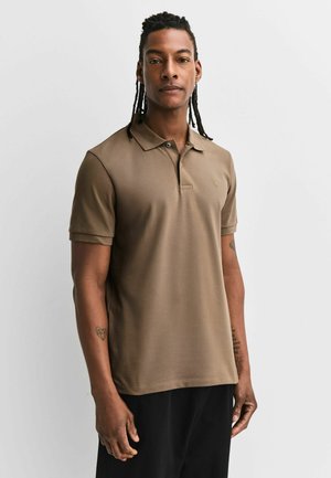 Bărbat cu păr împletit, purtând un tricou polo maro cu mâneci scurte și pantaloni negri, stând în fața unui fundal simplu și deschis la culoare.