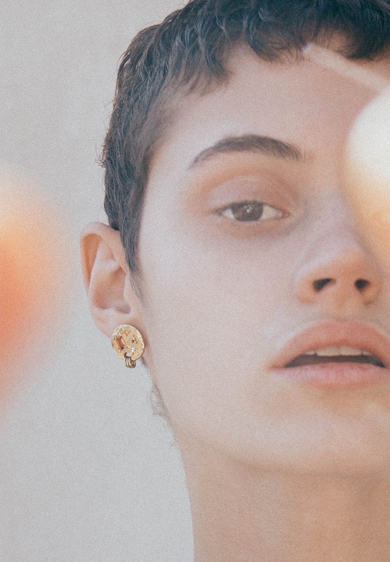 Kuldne kõrvarõngas, millel on keeruline geomeetriline kujundus, millel on kaunistuseks väike kalliskivi. Nahatoon on heledam.