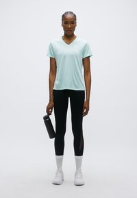 Αθλητική κοντομάνικη μπλούζα σε ανοιχτό μέντα, σε συνδυασμό με μαύρο κολάν με διακοσμητικές τομές από δίχτυ, λευκές κάλτσες και λευκά αθλητικά παπούτσια. Κρατά ένα μαύρο μπουκάλι νερού.