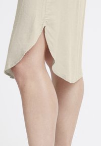 Jupe beige clair en tissu texturé, dotée d'une fente sur le côté et d'un ourlet arrondi, mettant en valeur un design doux et fluide.