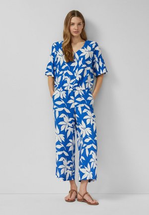 Frau steht vor einer grauen Wand, trägt ein blau-weiß geblümtes Kurzarmoberteil und passende, weit geschnittene Hosen mit beigen Sandalen.