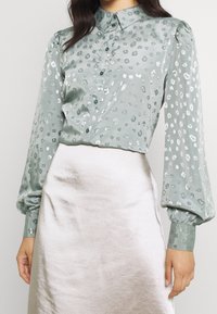 Lys grøn satinbluse med en subtil leopardprint, knappeskråkrave og pufærmer. Sammen med en beige satinneath.