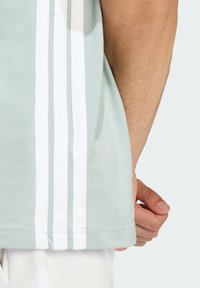 Světle zelené bavlněné tričko s třemi vertikálními bílými pruhy po straně, s kulatým výstřihem a krátkými rukávy.