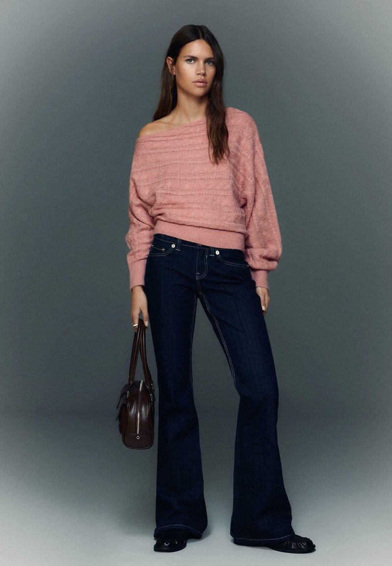 Lyserød off-shoulder strikket sweater med teksturerede detaljer, kombineret med mørkeblå udvidede jeans og en brun håndtaske. Enkle, rene linjer.