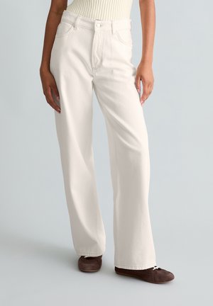 Personne portant un jean taille haute à jambes larges de couleur blanc cassé et des chaussures décontractées marron, debout devant un fond uni.