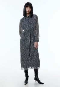 Langes schwarzes Kleid mit weißem Blumenmuster, durchsichtigem Stoff, Knopfleiste vorne und gebundenem Taillenbereich. Kombiniert mit glänzenden schwarzen Stiefeln bis zum Knie.