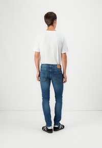Homme debout, de dos, portant un t-shirt blanc uni, un jean bleu slim et des baskets noir et blanc sur un fond blanc uni.