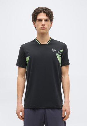 Νεαρός άνδρας με κοντά σγουρά μαλλιά, φορώντας μαύρο αθλητικό πουκάμισο με πράσινες πλαϊνές λεπτομέρειες με μοτίβο και λογότυπο στο στήθος, στέκεται μπροστά σε λευκό φόντο.