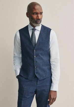 Uomo con barba grigia che indossa un gilet a quadri blu, pantaloni abbinati, camicia bianca e cravatta scura, in piedi con una mano in tasca.