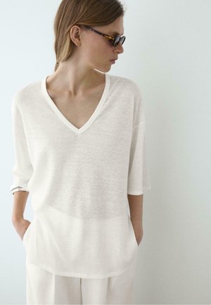 Kvinde iført lysebeige løst top med V-hals og matchende vidde bukser, med skilpaddemønstrede solbriller, hænderne i lommerne, foran ensfarvet baggrund.