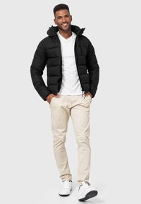 Schwarze Daunenjacke mit durchgehendem Reißverschluss und Kapuze, kombiniert mit einem weißen T-Shirt und beigen Hosen. Das Model trägt weiße Sneakers.
