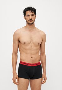 Boxers noirs avec une ceinture élastique rouge portant le logo "HUGO". Le tissu est lisse, s'ajuste étroitement au corps et offre une couverture minimale.
