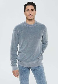 Sweat à mailles texturées grises avec un col rond côtelé et des poignets, associé à un jean bleu clair. Le tissu semble doux et confortable.