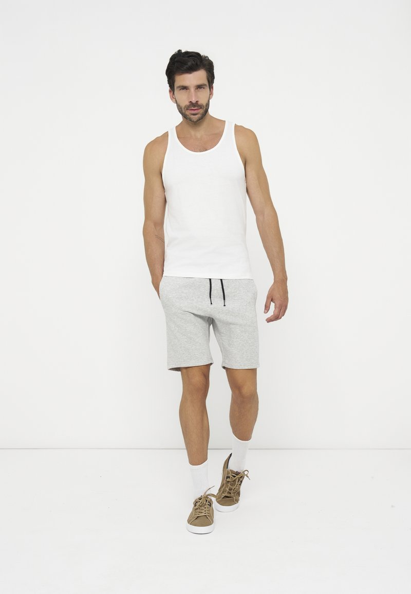 Mann, der geht, trägt ein weißes ärmelloses Shirt, hellgraue Shorts, weiße Socken und braune Sneakers vor einem schlichten weißen Hintergrund.