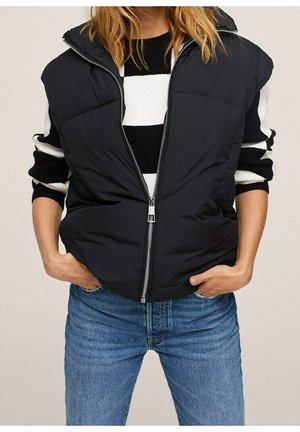 Kvinde iført en sort vatteret vest over en sort-hvid stribet sweater med blå jeans, hænderne i lommerne, stående mod en neutral baggrund.