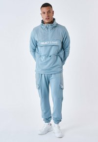 Ljusblå träningsdräkt i slätt material med en framficka, huva och elastiska muddarna. Vit logotyp tryckt på bröstet och byxorna.