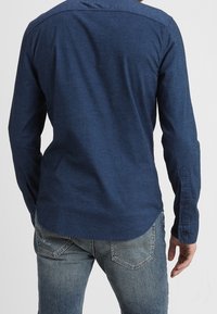 Modrá košile s dlouhým rukávem vyrobená z texturované látky, s kulatým lemem a knoflíkovými manžetami, vystavená zezadu.