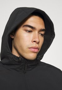 Veste à capuche noire avec une texture lisse, dotée de cordons réglables au niveau du cou et d'une fermeture éclair, affichant un design minimaliste.