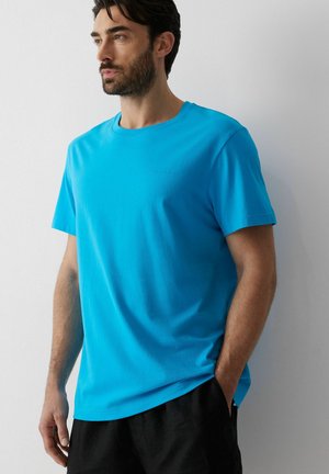 Mężczyzna o ciemnych włosach i brodzie, ubrany w jaskrawo niebieską koszulkę z krótkim rękawem i czarne spodnie, stojący przy prostym białym murze.