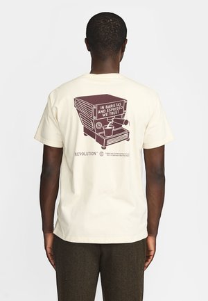 Mand iført beige t-shirt med brun espressomaskine grafik og teksten "In baristas and espresso we trust" på bagsiden.
