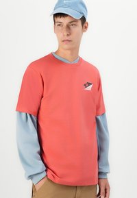 Young man wearing a coral red short-sleeve shirt over a light blue long-sleeve shirt and a light blue Nike cap, standing with one hand in pocket.